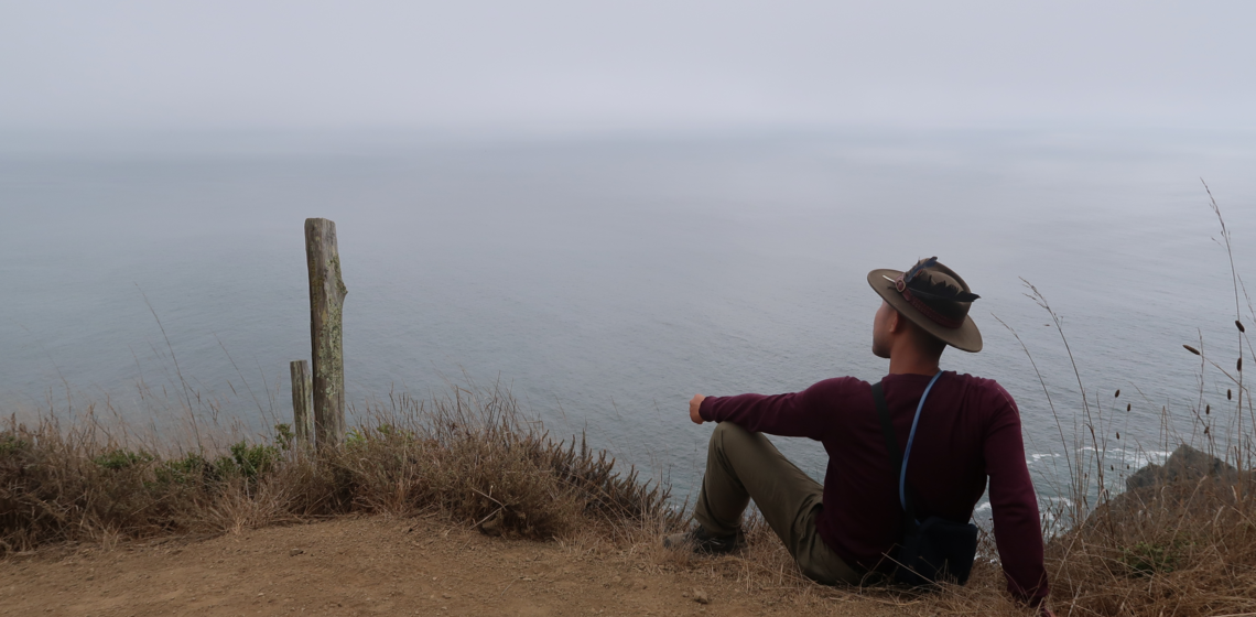 Student sitting on the edge of a mountain overlooking the graying sea