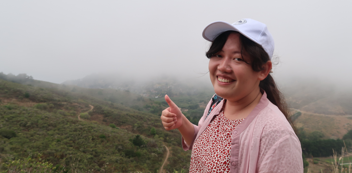 Student endorses the hike with a smile and a big thumbs up, standing on top the mountain overlooking the valleys below